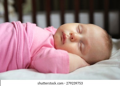 A Sweet One Month Old Newborn Baby Girl Is Sleeping On Her Back In Her Crib, Swaddled In A Pink Blanket.