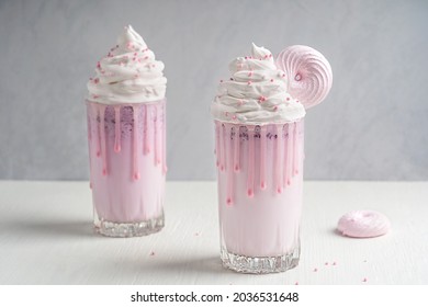 Sweet organic milkshake made of blended milk with icecream and berries decorated with topping of whipped cream and pink round meringue served in two drinking glasses on white wooden table. Horizontal