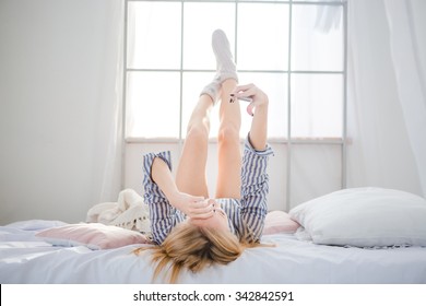 Sweet Relaxed Blonde Young Woman Lying On Bed With Legs Raised Up And Using Cell Phone