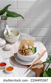 Sweet And Savory Chicken Noodle With Bean Sprout, Meatballs And Crunchy Cracker Served On White Bowl. Cafe Table Corner Situation With Some Indoor Plant. 