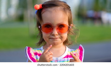 Sweet, Shy Toddler Baby Girl Wears Bright Striped Summer Bodysuitin And Round Orange Spectacles Outdoors. Close Up Portrait Little Kid Child In Summer Public Park