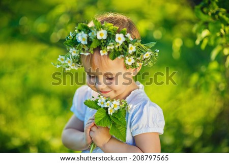 A sweet smiling little girl with a wreath of wild strawberry flowers in a white dress holding a twig of this plant in a green forest in a sunny summer day closeup