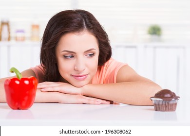 Sweet Temptation. Thoughtful Young Woman Choosing What To Eat While Leaning At The Kitchen Table With Chocolate Muffin And Fresh Pepper Laying On It