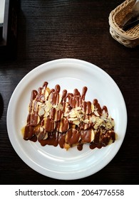 Sweet Thin Pancake (crepe) Covered With Sliced Banana, Almond And Served With Hot Fudge On White Plate In Background Of Dark Wooden Table And Tableware Aside In Restaurant. Warm Chocolate Dessert