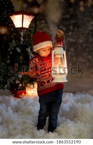 Sweet toddler blond boy with red sweater, holding lantern in the forest, christmas time, snowing