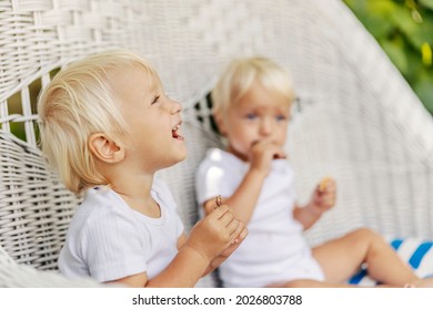 Sweet Toddler Twins, Funny Family Moment. The Cute Babies In The White Bodysuits Enjoy Eating Cookies And While Sitting On A Large Canvas Chair. Twins With Blue Eyes And Blonde Hair Enjoying The Sun