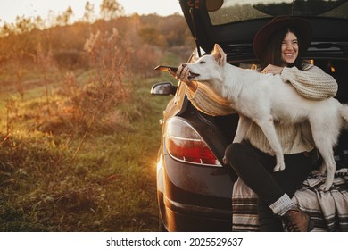 Sweet white dog biting phone while happy stylish woman taking selfie photo in car trunk in sunset light in field, funny moment. Autumn road trip with pet and travel. Young hipster female using phone - Shutterstock ID 2025529637