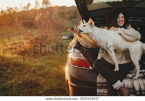 Sweet white dog biting phone while happy stylish\
woman taking selfie photo in car trunk in sunset light in field,\
funny moment. Autumn road trip with pet and travel. Young hipster\
female using phone