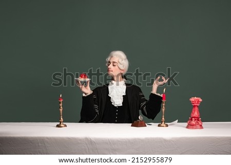 Sweets, cakes. Portrait of young elegant man in peruke and vintage jacket sitting at table isolated on dark green background. Retro style, comparison of eras concept.