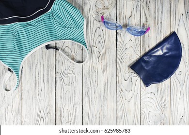 Swim Items (swimsuit, Swimming Goggles, Swimming Cap) On The Wooden Background. Top View