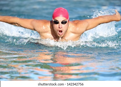 Swimmer athletic man swimming butterfly in a swimming cap approaching the camera rising up out of the water. Male athlete fitness model training outside.