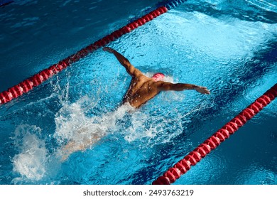 Swimmer in pink cap executes powerful butterfly stroke, his muscular form cutting through blue water, creating dynamic splash between red lane dividers. Concept of sport, preparation to competition - Powered by Shutterstock