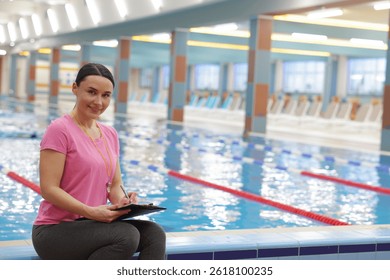 Swimming coach taking notes while sitting on edge of indoor pool. Space for text