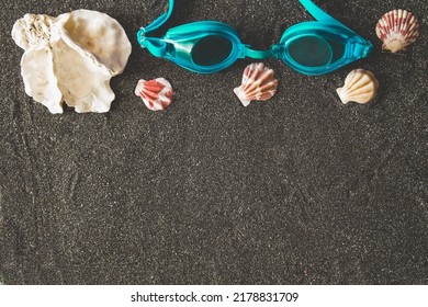 Swimming Goggles And Assorted Seashells On Black Volcanic Sand Background. Top View. Space For Text. Tropical Vacation Concept