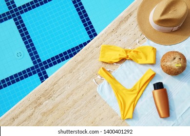 Swimming pool accessories concept. Top view of beach items on deck, bright yellow bikini bathing suit, hipster straw hat, blue mat & sunscreen skincare cream. Colorful beach wear. Flat lay, copy space