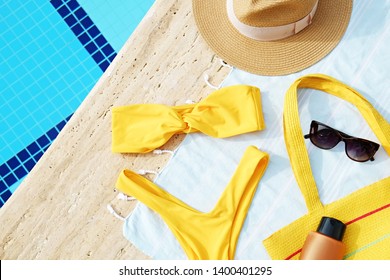 Swimming pool accessories concept. Top view of beach items on deck, bright yellow bikini bathing suit, hipster straw hat, blue mat & sunscreen skincare cream. Colorful beach wear. Flat lay, copy space