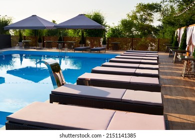 Swimming Pool Area With Deck Chairs And Sun Umbrellas Along The Poolside In Luxury Resort. Beach Beds Surrounded With Greenery And Blue Water. Tranquility And Relax Atmosphere On Vacation.