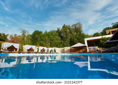 Swimming Pool Area With Gazebos, Deck Chairs And Sun Umbrellas Along The Poolside In Luxury Resort. Summer Houses Surrounded With Greenery And Blue Water. Tranquility And Relax Atmosphere On Vacation.