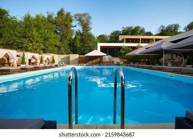 Swimming Pool Area With Ladder, Gazebos, Deck Chairs And Sun Umbrellas Along The Poolside In Luxury Resort. Summer Houses Surrounded With Greenery. Tranquility And Relax Atmosphere On Vacation.