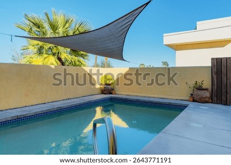 Swimming pool in backyard with triangle sun shade sail above. Yellow wall and palm trees in the background.