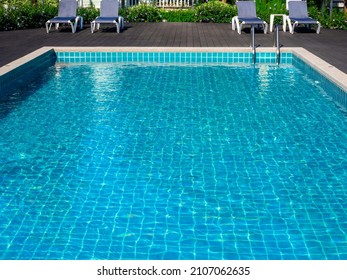 Swimming Pool With Clean Clear Water, Nobody. Square Shaped Pool With Blue Tiles With Grab Bars Ladder And Sunbeds On The Wood Decks, No People. Water Surface. Overhead View.  Summer Background.