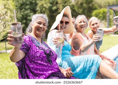 At swimming pool, diverse senior female friends enjoying drinks, smiling at camera. Biracial woman with purple background, Asian with blue, two Caucasians with pink and teal, sharing laughter under - Powered by Shutterstock