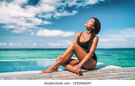 Swimsuit model beauty skincare suntan Asian woman posing sunbathing tanning in black one piece bathing suit. Beautiful girl relaxing at overwater infinity pool luxury resort. Spa, wellness, laser.