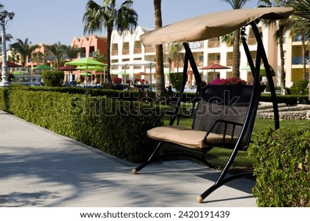 Swing bench with shelter from the sun stands along a hedge on a pedestrian road in a park for a walk on a sunny day