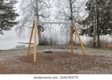 A swing set made of yellow and grey materials is positioned in a frost-covered playground near a misty lake. The atmosphere is calm with surrounding trees. - Powered by Shutterstock