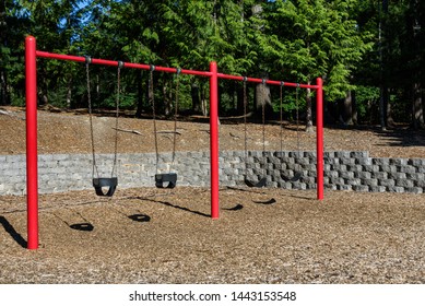 Swing set on a sunny day in a park playground, four classic black rubber swings with red posts, wood chips below and woodland behind
