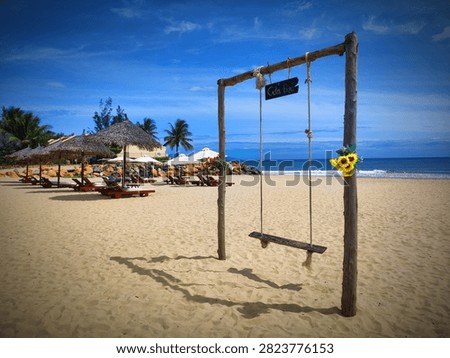 A swing set stands on the sandy beach with a view of the ocean. Palm trees and lounge chairs are visible nearby under clear blue skies. The scene shows leisure and relaxation.