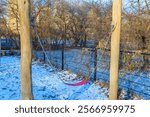 A swingset sits in a snowy park surrounded by leafless trees and a river, evoking a serene winter atmosphere.