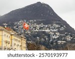 Swiss flag waving, with the city of Lugano and Monte Bre forming the backdrop in Switzerland