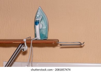 An Switched Off Iron With A Transparent Water Tank Stands On An Empty Ironing Board Against The Wall