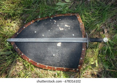 Sword lying on a triangular shield, on the ground in a tall green grass. The lost weapons after the battle, the battlefield. Medieval weapons of the knight.