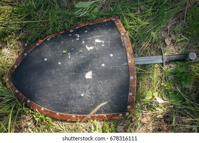 Sword lying under a triangular shield, on the ground in a tall green grass. The lost weapons after the battle, the battlefield. Medieval weapons of the knight.