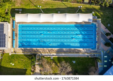 Sydney, Australia: Jun 26 2020: Overhead Top Down Aerial Shot Of An Outdoor Olympic Size, Six Lane, Swimming Pool In Afternoon Light, With People Swimming Laps