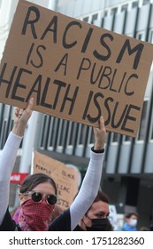 Sydney, NSW / Australia - June 6 2020: Black Lives Matter. Young Woman Marching With The Protesting Crowd Wearing Sunglasses And A Bandana. Holding A Sign Reading 'racism Is A Public Health Issue'