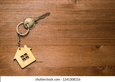 Symbol Of A Bamboo House With A Metal Key On A Brown Vintage Wooden Background. Lighting Gradient. The Concept Of Selling Real Estate With The Transfer Of Ownership.