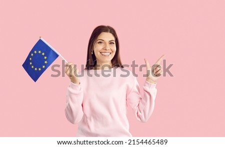 Symbol of Europe, EU association. Caucasian woman holding small flag with european union emblem isolated on pink background. Happy and smiling woman holding flag and pointing finger at copy space.
