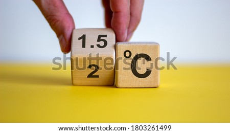 Symbol for limiting global warming. Male hand turnee a cube and changes the expression '2 C' to '1.5 C', or vice versa. Concept. Beautiful yellow table, white background, copy space.