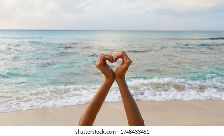 Symbol Of Love For Nature. Female Hands Near The Ocean On The Beach Show A Heart Symbol On A Background Of Turquoise Clear Water Of The Sea