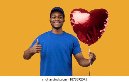 Symbol Of Love. Smiling Black Delivery Man In Blue Uniform And Cap Holding Red Helium Party Balloon In Heart Shape And Gesturing Thumb Up Sign Isolated On Yellow Orange Background. Valentine's Day