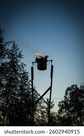 A symbolic Midsummer fire burns in an elevated metal cauldron, standing high against the evening sky. Decorated with oak leaves, the flame represents purification, renewal, and the Baltic solstice.