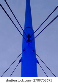 A symmetrical close-up view of a cable-stayed bridge tower illuminated in blue light, with tension cables radiating outward against a soft evening sky. Modern engineering and design highlighted.