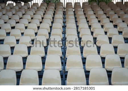 Symmetrical full frame view of many empty beige plastic seats in rows on stadium grandstand before sporting event or concert during outdoor day.