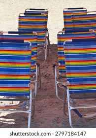 A symmetrical, high angle view of empty rainbow striped folding beach chairs arranged in rows on a sandy shore, evoking a colorful and nostalic summer vacation aesthetic.