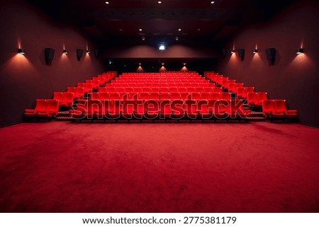 Symmetrical interior view of a luxurious empty cinema hall with a wide red carpet foreground. The rows of vibrant red velvet seats are arranged in a tiered layout, framed by dark walls with elegant warm-toned sconce lighting. A perfect representation of the cinematic experience, premiere night, or a vacant theater venue ready for an event.