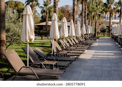 Symmetrical row of empty textile mesh sunloungers with folded canvas patio umbrellas on a modern paved terrace walkway of a resort hotel