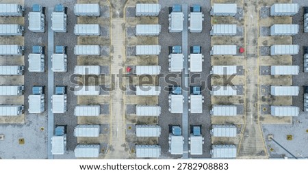 Symmetrical rows of white containerized battery units on gravel pads, intersected by paved service lanes with directional markings, under diffuse daylight, Aerial view of energy storage power station